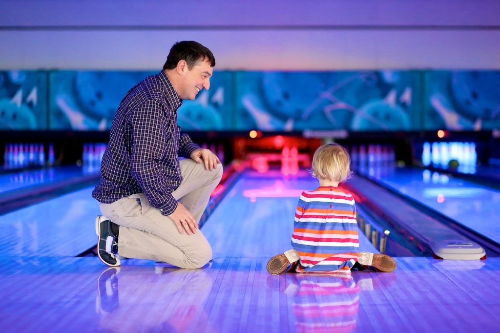 Happy family of two, active father with little daughter, adorable blonde toddler girl, playing bowling together enjoying weekend leisure activity