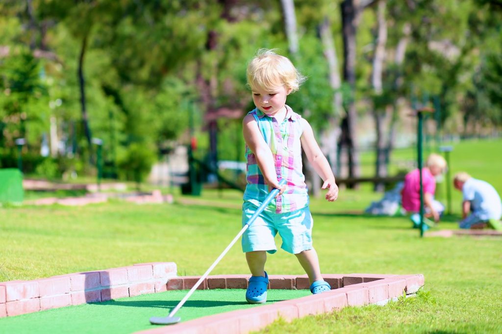 Group of happy active children, two brothers, teenage boys, and their little sister, cute toddler girl, playing miniature golf enjoying sunny summer vacation day outdoors in the park